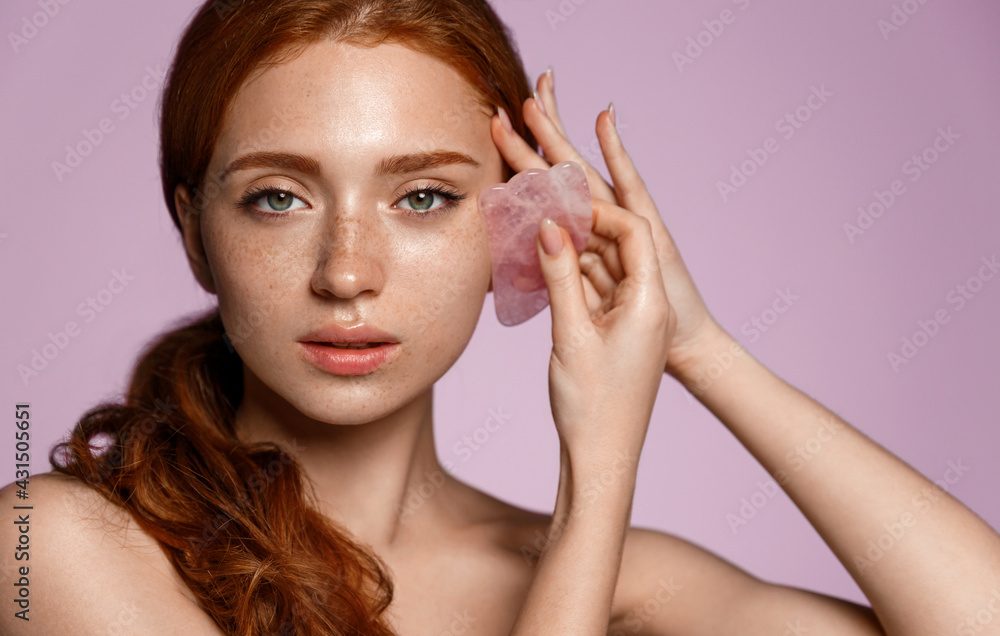 Redhead girl uses gua sha quarz lifting tool on face. Woman self massage with jade gouache scraper, regulating blood flow with tcm spa treatment, apply skincare cream serum, reduce facial tension - obrazy, fototapety, plakaty 