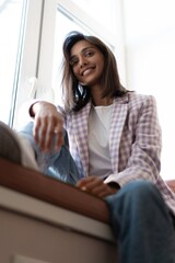 Canvas Print - Young Indian mixed-race woman dressed casually sitting on the window sill at home office