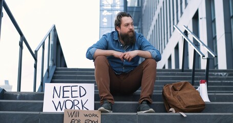 caucasian young sad and depressed man in despair sitting on stairs in city with posters need work an