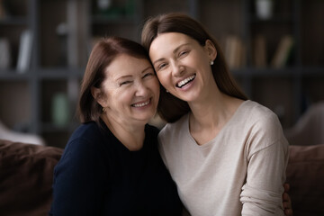 Wall Mural - Portrait of happy two generations of women pose at home relax together. Smiling elderly Caucasian mother and adult grownup daughter rest on sofa, enjoy family weekend time. Unity concept.