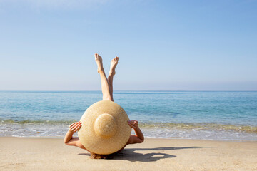 Young woman lying on the beach with legs pointing up to the sky covering her body with broad brim straw hat. Female sunbathing on the shore of atlantic ocean. Close up, copy space, back view.