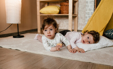 two little twin girls play lying on the floor with cookies in their hands near the yellow teepee.