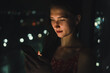 © yaroslav1986 - Portrait of a beautiful young woman in a red dress and with red lipstick on her lips standing near the window at night holding a mobile phone near her face, soft light from the phone falls on her face