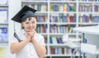 Smiling girl with Down Syndrome wearing a graduation cap stands at a library. Empty space for text. Education for disabled children concept