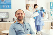 © DC Studio - Man patient looking into camera waiting for radiography results sitting on dental chair in hospital stomatology clinic. In background dentists team looking at tooth x-ray preparing for surgery