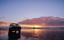 White Ute Parked On The Beach And Lone Person Standing By The Water At Sunset