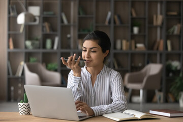 Indian businesswoman recording voice audio message for social network, sitting at desk with laptop, holding smartphone, young woman chatting online or speakerphone, activating digital assistant