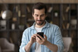 © fizkes - Close up satisfied man using phone, looking at screen, standing at home, positive young male holding smartphone, chatting in social network with friends or shopping online, enjoying leisure time