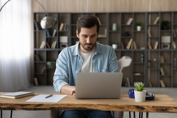 Confident businessman working on laptop online, typing, writing business email or financial report, sitting at wooden desk at home, focused young man searching information, educational course