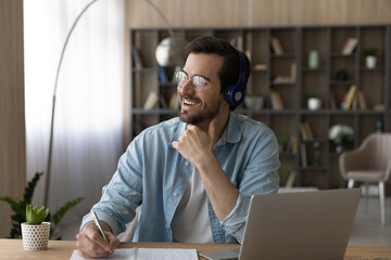 Wall Mural - Dreamy excited man in headphones and glasses distracted from online studying, looking to aside, sitting at desk with laptop, visualizing opportunities, happy student taking notes, watching webinar