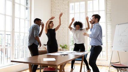 Overjoyed millennial diverse mixed race business people dancing in modern office, celebrating shared corporate success. Emotional happy multiracial employees having fun, relaxing together at workplace