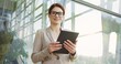 © VAKSMANV - Portrait of the Caucasian beautiful businesswoman, taping on the tablet computer, smiling to the camera and posing on the glass building background. Outdoors