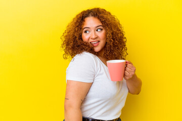 Young latin curvy woman holding a cup isolated on yellow background looks aside smiling, cheerful and pleasant.