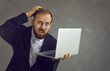 © Studio Romantic - Confused bearded businessman holding a laptop and scratching his head while standing on a gray background. Middle-aged man with a frustrated expression gets bad or negative news. Banner.