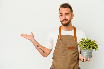 Young gardener tattooed caucasian man holding a plant isolated on white background showing a copy space on a palm and holding another hand on waist.