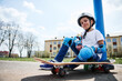 Bottom view of smiling boy in protective gear of skateboarder sitting on skateboard and looking at camera