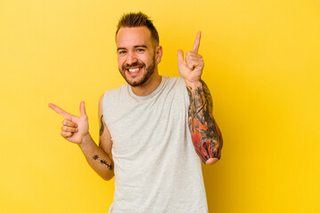 Young tattooed caucasian man isolated on yellow background pointing to different copy spaces, choosing one of them, showing with finger.