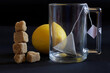 A glass transparent mug with a pyramid tea bag, fresh yellow lemon and a tower of brown cane unrefined sugar cubes. Dark background
