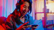 © tirachard - Happy asia girl gamer wear headphone competition video game online with smartphone excited talk with friend sit on couch in colorful neon lights living room at home, Home quarantine activity concept.