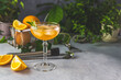 Champagne coupe glass of refreshing orange cocktail with ice served on gray table surface surround of orange fruit and different green plants