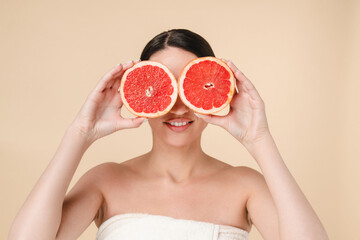 Wall Mural - Young attractive caucasian woman holding grapefruits near her face isolated over beige background. White woman in spa towel waiting for spa and body care procedures. Pampering, moisturizing effect