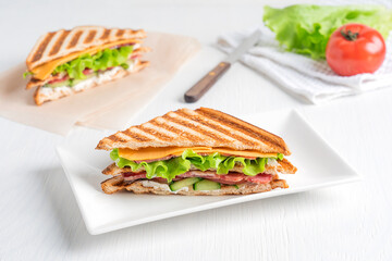 Poster - Homemade healthy sandwich made of cucumber, slice of meat and cheese, lettuce and tomatoes between slices of grilled bread served on plate on white wooden background with ingredients. Horizontal