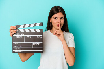 Young caucasian woman holding a clapperboard isolated on blue background keeping a secret or asking for silence.