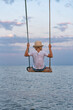 © somemeans - Boy in straw hat riding on swing over the water. Swing against the sky and the sea