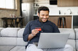 © Vadim Pastuh - Happy indian guy holding credit card and paying online, using laptop computer for shopping online, transferring money, mixed-race man sits on the couch and input banking card data for purchasing