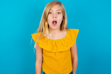 Wall Mural - Oh my God. Surprised Caucasian kid girl wearing yellow T-shirt against blue wall stares at camera with shocked expression exclaims with unexpectedness,