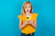 © Roquillo - Caucasian kid girl wearing yellow T-shirt against blue wall using mobile phone chatting free time .