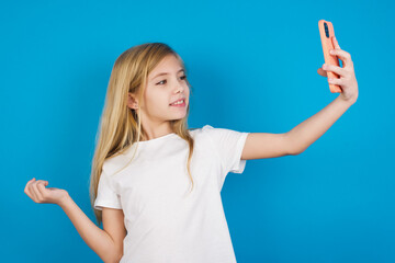 Wall Mural - Portrait of a beautiful Caucasian little girl wearing white T-shirt over blue background taking a selfie to send it to friends and followers or post it on his social media.