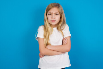 Wall Mural - Gloomy dissatisfied beautiful Caucasian little girl wearing white T-shirt over blue background looks with miserable expression at camera from under forehead, makes unhappy grimace