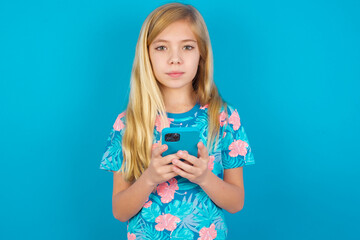 Portrait of serious confident Caucasian kid girl wearing hawaiian T-shirt against blue wall  holding phone in two hands