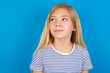 © Roquillo - Caucasian kid girl wearing striped shirt ​against blue wall looking aside into empty space thoughtful