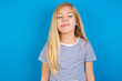 © Roquillo - Caucasian kid girl wearing striped shirt ​against blue wall showing grimace face crossing eyes and showing tongue. Being funny and crazy