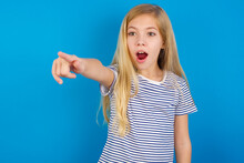 Caucasian Kid Girl Wearing Striped Shirt ​against Blue Wall  Pointing With Finger Surprised Ahead, Open Mouth Amazed Expression, Something On The Front.