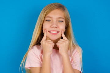 Happy Caucasian kid girl wearing pink shirt against blue wall with toothy smile, keeps index fingers near mouth, fingers pointing and forcing cheerful smile