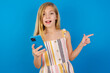 © Roquillo - Astonished Caucasian kid girl wearing dress against blue wall holding her telephone and pointing with finger aside at empty copy space