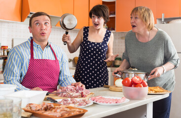 Irritated woman with elderly mother scolding her husband cutting meat for family dinner in home kitchen. Family conflict