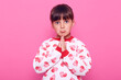 Sweet little girl with dark hair looks at camera with a pleading expression on her face, keeping palms together, praying, wearing a stylish sweater, isolated over pink background.