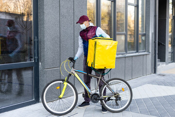 Wall Mural - Young courier in medical mask delivering food with yellow thermal backpack, riding a bicycle in the city. Food delivery service concept