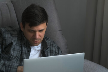 Smiling man is typing a message and reading on laptop lying on sofa at home. Freelance, working, having a job concept. Friendly chatting on e-mail. Using online dating computer app alone at home.