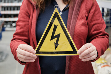 Woman holds yellow triangular sign with lightning bolt painted on it. The sign warns of high electrical voltage. Concept of warning signs and mandatory compliance with occupational safety regulations