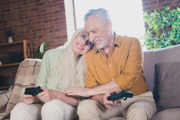 Wall Mural - Photo of happy affectionate old man and woman family sit sofa play game joystick indoors inside house home