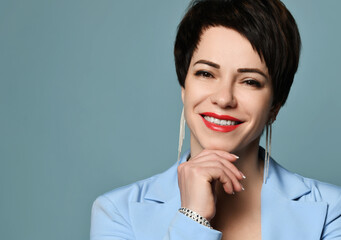 Wall Mural - Portrait of beautiful smiling short haired brunette woman in blue business smart casual suit and earrings looking at camera over blue background. Stylish business female wear and fashion concept