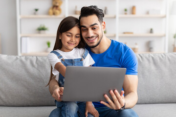 Wall Mural - Smiling dad and daughter sitting on couch, using laptop