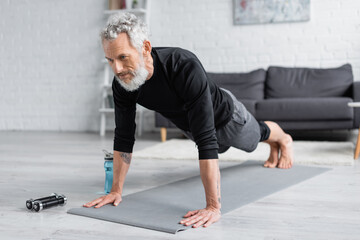 tattooed man with grey hair doing plank on fitness mat near dumbbells and sports bottle in living room.