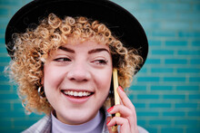 Smiling Curly Haired Woman In Hat Looking Away While Talking On Smart Phone