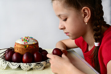 A cute girl sits at a table with a traditional Easter cake and colored eggs and holds a red egg in her hand. Festive treat at Easter